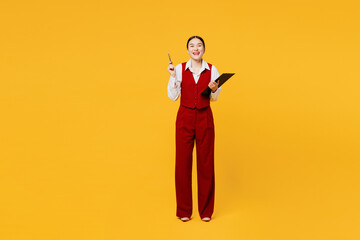 Full body young lawyer employee business woman of Asian ethnicity wear red vest shirt work at office hold clipboard paper documents point finger up isolated on plain yellow background. Career concept.