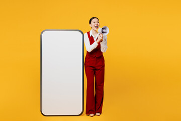 Full body young lawyer employee business woman of Asian ethnicity wear formal red vest shirt work at office big blank screen mobile cell phone scream in megaphone isolated on plain yellow background.