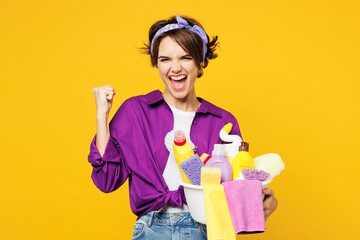 Young excited woman wear purple shirt hold basin with detergent bottles do housework tidy up doing winner gesture celebrate isolated on plain yellow background studio portrait. Housekeeping concept. - obrazy, fototapety, plakaty