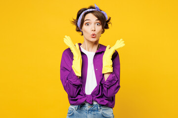 Young surprised shocked fun woman wear purple shirt rubber gloves while doing housework tidy up spreading hands look camera isolated on plain yellow background studio portrait. Housekeeping concept.