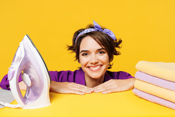 Close up young smiling happy woman wear purple shirt casual do housework tidy up ironing clean clothes on board look camera isolated on plain yellow background studio portrait. Housekeeping concept.