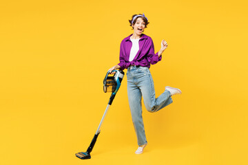 Full body happy young woman wear purple shirt casual clothes do housework tidy up hold vacuum cleaner raise up leg look camera isolated on plain yellow background studio portrait Housekeeping concept