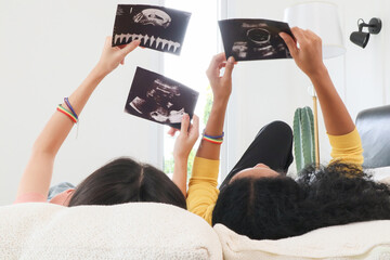 A multiracial lesbian couple lying watching ultrasound images, two lesbians feeling excited about having their first baby by IVF (In vitro fertilization) process. Concept of pregnancy and motherhood - obrazy, fototapety, plakaty