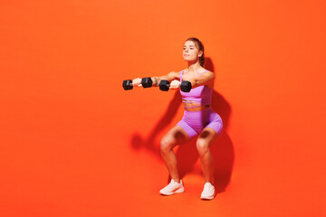 Full body young fitness trainer instructor woman sportsman wear top shorts purple clothes train in home gym hold dumbbells do squats isolated on plain orange background. Workout sport fit abs concept.