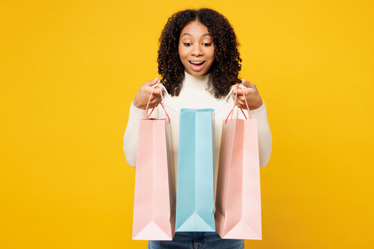 Little surprised happy kid teen girl wearing white casual clothes hold open look at paper package bags after shopping isolated on plain yellow background. Black Friday sale buy day lifestyle concept.