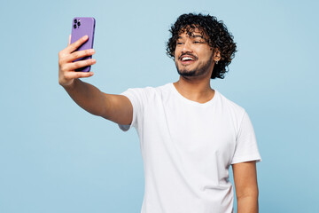 Young happy Indian man he wear white t-shirt casual clothes doing selfie shot on mobile cell phone post photo on social network isolated on plain pastel light blue cyan background. Lifestyle concept.