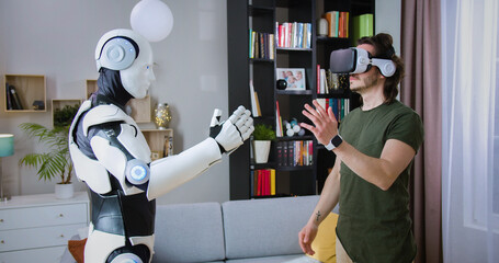 Focused man in virtual reality headset examining robot's movements in bright living room. Humanoid...