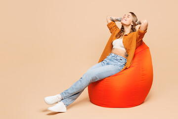 Full body happy young woman she wear orange shirt casual clothes sit in bag chair hold hands behind neck close eyes isolated on plain pastel light beige background studio portrait. Lifestyle concept. - obrazy, fototapety, plakaty
