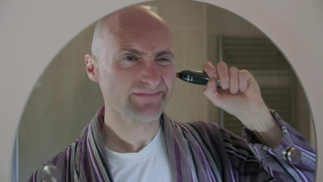 CU 50fps. Bald middle aged man, shaving his nose and ear hair in the bathroom mirror, with electric shaver. Funny facial expression.