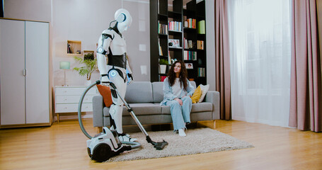 Happy young woman with curly hairstyle enjoying her leisure time while robot doing work in...