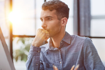 Young pensive coworker working at sunny work place loft while sitting at the wooden table