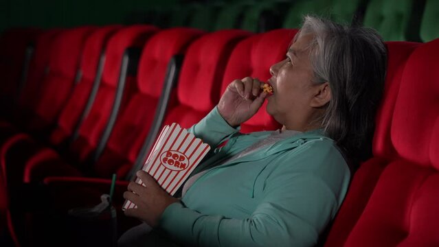 Asian elderly women eating popcorn and watching exciting or scary movie at cinema. alone asia senior old female holding a bucket of popcorn in theater