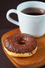 Donut with chocolate icing next to a white cup of tea on a wooden board on a dark background