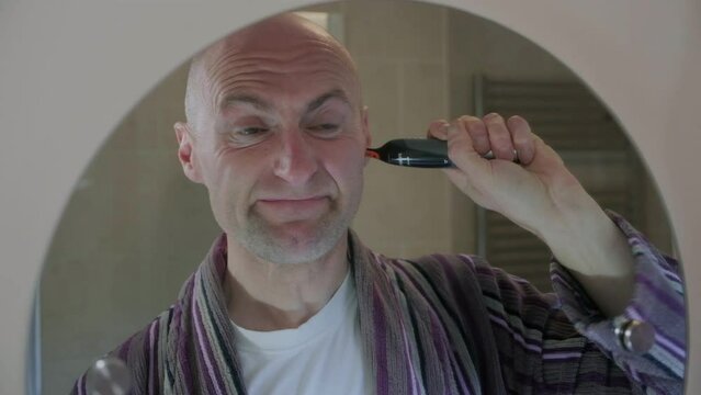 CU 50fps. Bald middle aged man, shaving his nose and ear hair in the bathroom mirror, with electric shaver. Funny facial expression.