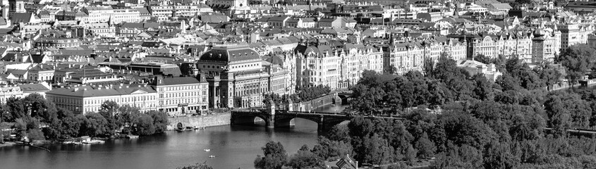 A view overlooking the National Theatre in Prague, with the Vltava River and a bridge in the foreground surrounded by trees and architecture. Prague, Czechia. Black and white photography.