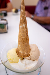 An appetizing cup of ice cream with three yellow, white and beige scoops. An upside down cone to garnish it. In the blurred background are the people who ordered it.