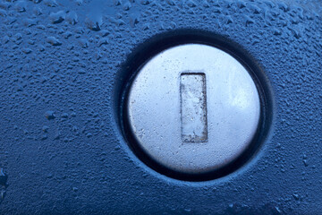 A close-up view of a car door lock, adorned with delicate dew drops that glisten in the light. The metallic surface of the lock contrasts with the cool blue and gray tones of the car’s body