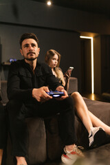 Handsome man with moustache holds joystick sitting near girlfriend with smartphone. Relaxed couple spends time in club together