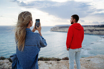 Woman uses her smartphone to photograph a man in a red hoodie standing on the edge of a cliff with...