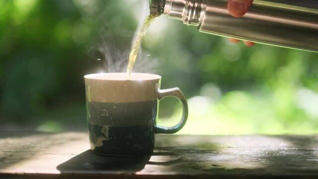 Cup of hot tea poured from a thermos on the railing of the terrace against the backdrop of a green garden and sunlight, 4k video footage