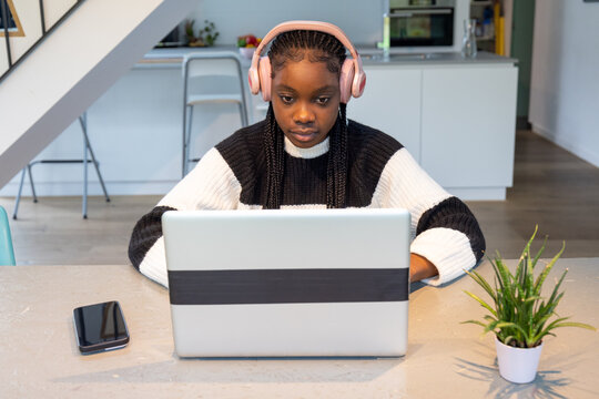 A young African American girl is deeply engrossed in her studies while wearing pink headphones, using a laptop in a bright, modern kitchen. A smartphone rests beside her, and a green plant adds a