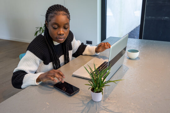 This image features a young African American woman in a black and white sweater, seamlessly multitasking in a minimalist workspace. She operates a smartphone and a laptop simultaneously, suggesting