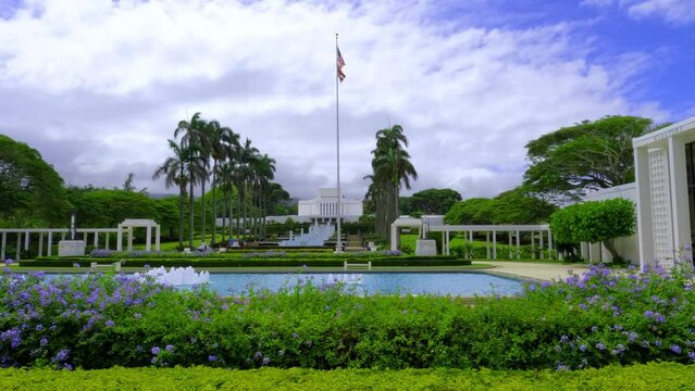 American flag flutters in the wind above the park near Laie Hawaii Temple. A view of Laie Hawaii Temple park with fountain pools and the state flag flipping in the wind. 