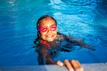 A young girl with a beaming smile wears pink goggles while swimming in a sparkling blue pool,...