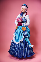 Fototapeta premium cute stylish female cosplayer in vivid attire holding pink flowers and looking away on pink backdrop
