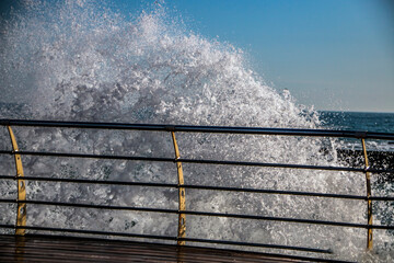 Huge waves roll onto a wooden pier with railings against the backdrop of a clear blue sky and sea...