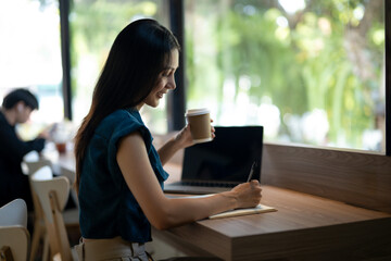 Smiling young woman engrossed in writing notes in a notebook in a café with a takeaway coffee cup...