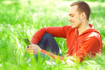 Naklejka na ściany i meble Gadget lover concept. Portrait of smiling young man in casual clothing reading e-book, sitting in green grass in the park. Great white shiny smile. Close up. Copy-space. Sunny weather. Outdoor shot