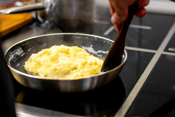 person is cooking scrambled eggs in a pan on a stovetop, stirring with a wooden spatula. The steam...