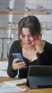 Vertical video. Young woman sit in the kitchen experiences severe pain in neck rubbing to relieve muscle tension massaging neck while using smartphone in kitchen at home	
