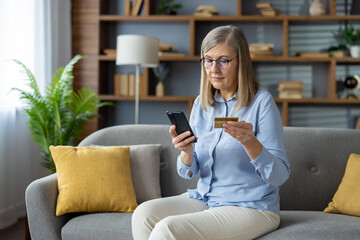 Mature woman sitting on couch at home, using smartphone and holding credit card for online shopping.