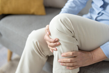 Close-up image of a mature woman sitting on a couch and holding her knee, depicting pain or discomfort in the joint. - obrazy, fototapety, plakaty