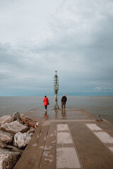 Senior couple having a romantic walk on the pier in a Riviera Romagnola beach location, bright red lady jacket, contrast with the dramatic sky in a winter afternoon. Winter sea.