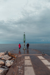 Senior couple having a romantic walk on the pier in a Riviera Romagnola beach location, bright red lady jacket, contrast with the dramatic sky in a winter afternoon. Winter sea.
