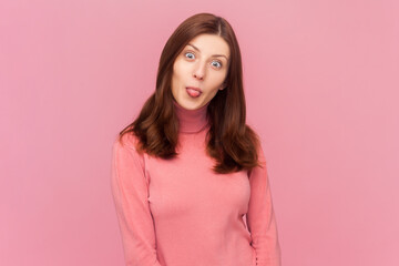 Funny woman with brown hair sticks out tongue and makes grimace, has fun mimics childish face, stays happy and positive, wearing rose turtleneck. Indoor studio shot isolated on pink background - obrazy, fototapety, plakaty