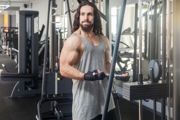 Fototapeta na wymiar Portrait of muscular attractive man with long curly hair training at gym lifting weights using simulator, doing exercises for biceps and triceps, looking away has workout for arms. Indoor shot.