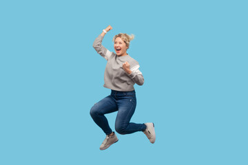 Obraz premium Full length portrait of overjoyed happy blonde woman jumping up with clenched fists celebrating her victory, wearing gray sweatshirt. Indoor studio shot isolated on blue background.