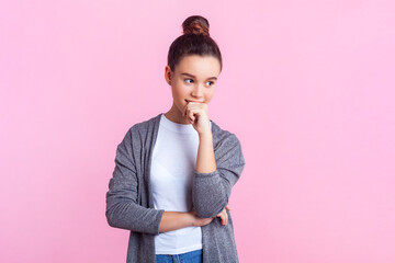 Portrait of pensive teenage girl with bun hairstyle in casual clothes standing holding chin...