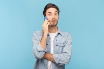 Portrait of surprised shocked amazed unshaven man talking on mobile phone, looking at camera with big eyes, hearing breaking news, wearing denim shirt. Indoor shot isolated on blue background.