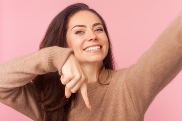 Pleasant looking woman with wavy hair, making selfie or broadcasting livestream, pointing down, asking to subscribe, wearing wearing brown pullover. Indoor studio shot isolated on pink background - Powered by Adobe