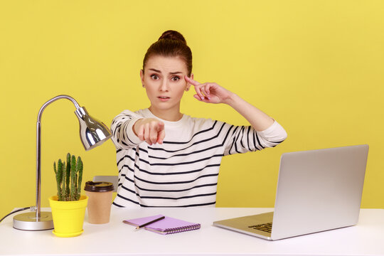 Woman with sarcastic look sitting at workplace and making stupid gesture, pointing to camera, teasing and accusing senseless dumb mind. Indoor studio studio shot isolated on yellow background.