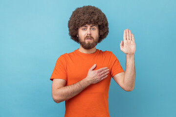 Portrait of man with Afro hairstyle wearing orange T-shirt holding hand on heart, juryman swearing...
