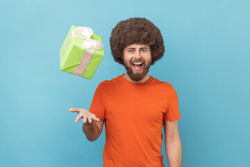 Portrait of happy funny optimistic man with Afro hairstyle wearing orange T-shirt throwing present...