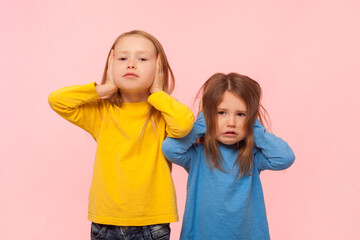 Portrait of two serious annoyed little girls covering ears with palms, do not want to hear friend...