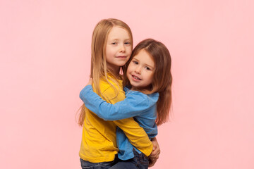 Portrait of two loving friendly little girls sisters standing hugging each other, expressing support and love, looking at camera. Indoor studio shot isolated on pink background.