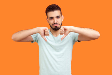 Portrait of displeased sad unhappy young bearded man wearing T-shirt standing showing thumbs down looking at camera with negative feelings. Indoor studio shot isolated on orange background. - obrazy, fototapety, plakaty